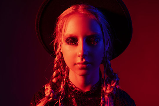 Conceptual portrait with the eyes closed of a beautiful teenage girl wearing a black dress and a black hat. It is artistically illuminated by red and blue light
