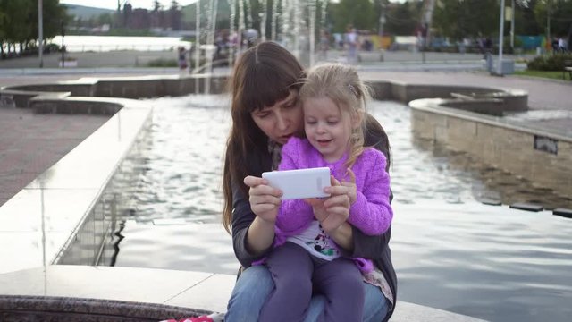 A little girl is using the application in a smartphone, her mother is holding a phone. The family sits on a bench in a public park on a cloudy day.