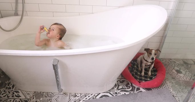 Adorable baby boy is sitting in water in bathtub with soap. Dog sits next to it on its bedding