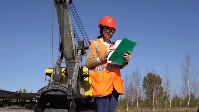 A young woman worker in sunglasses stands near a mining excavator, looking over project.