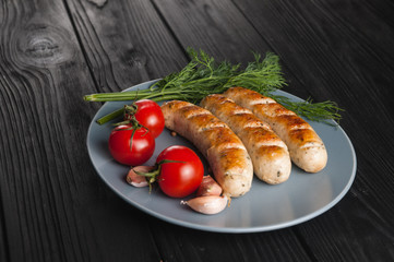 Three grilled sausages on a gray plate on a wooden black background with cherry tomatoes, herbs, garlic.