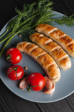 Three grilled sausages on a gray plate on a wooden black background with cherry tomatoes, herbs, garlic.