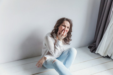 Happy teenage girl smiling. Young happy positive woman in white shirt sitting on floor in bright...