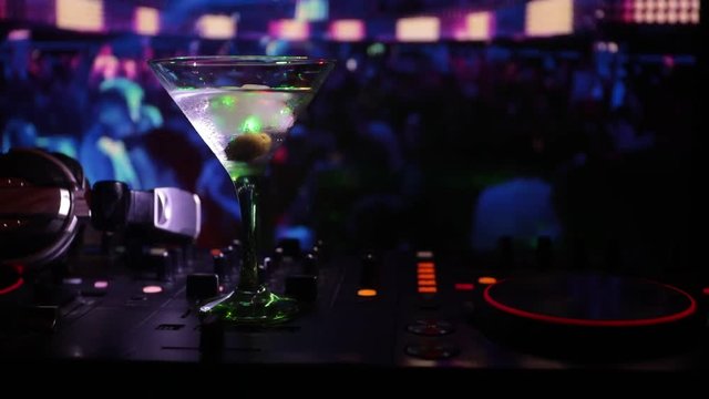 Glass with martini with olive inside on dj controller at nightclub. Dj Console with club drink at music party in nightclub with disco lights.