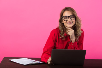 beautiful young girl in a red shirt and glasses near a laptop on a pink background