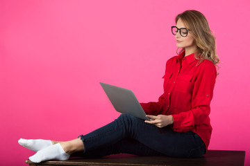 beautiful young girl with glasses in a red shirt sitting at the desk with a laptop on a pink background