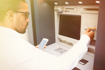 Human and technology. Dark skinned man using ATM. Black guy's hand inserting plastic bank card into...