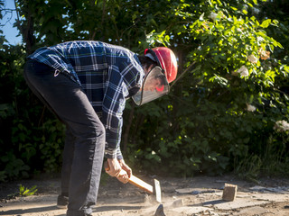 young male worker in protective mask crashing the stone with hammer outside on a summer day