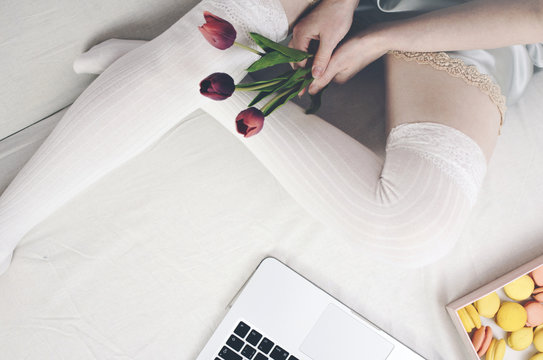 Female legs in white stockings on the bed with flowers in their hands and macaroons.