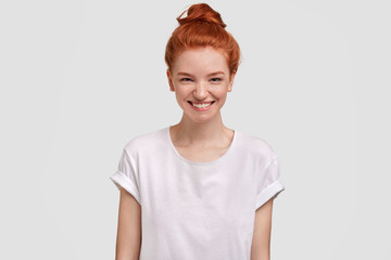 Positive young red haired cute female has freckled skin and joyful expression, rejoices success, dressed in casual t shirt, isolated over concrete wall. Ginger teenager satisfied with something.