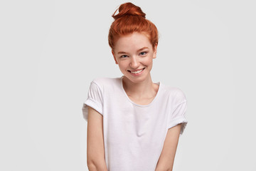 Portrait of happy red haired female being in good mood, wears casual clothing, has shy expression, recieves compliment, stands against white background. Isolated shot. People and skin care concept