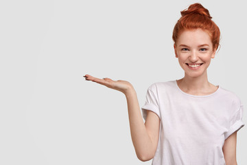 Lovely foxy female demonstrates boutique, keeps palm raised, stands against white background, has positive expression and freckled skin. Pointing at great place, where there is something interesting