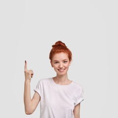 Delighted ginger girl indicates with fore finger upwards at blank copy space above her head, shows...
