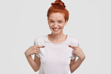 Candid shot of good looking female with red hair combed in bun, indicates at blank space of white...