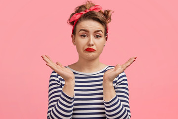 Portrait of puzzled hesitant young female model dressed in retro clothing, shrugs shoulders as doesn`t know what to wear on party, has doubtful expression, isolated on pink background. So what?