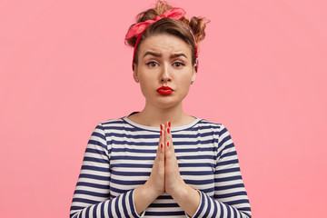 Portrait of good looking sorrowful female with pleading expression, keeps hands in praying gesture, asks for something, wears striped sailor jacket and stylish headband, has red painted lips