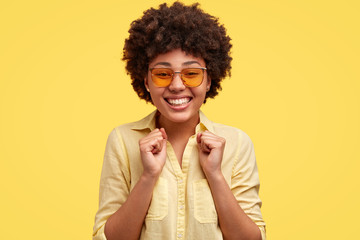 Fototapeta na wymiar Glad positive African American female watches football match, keeps hands in fists, shouts for her favourite team, wears stylish clothes, poses against yellow background. Woman winner with dark skin