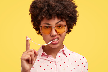 Photo of good looking young African American female has angry expression, stretches bubble gum,...