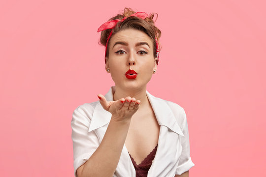 Attractive pin up girl with wonderful make up, blows air kiss at camera, wears retro clothing, flirts with man, poses against pink background. Gorgeous young lady has red lips sends kiss to you