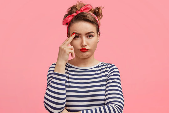 Bored tired thoughtful female has serious pensive expression, keeps fingers near eyebrows, has specific look, wears stylish red headband, stands against pink background. Retro style concept.