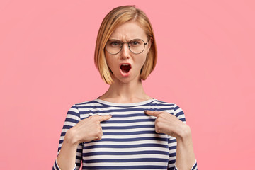 Grumpy annoyed angry female model wears casual sailor jacket and spectacles, indicates at blank...