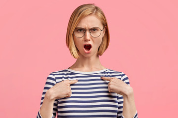 Grumpy annoyed angry female model wears casual sailor jacket and spectacles, indicates at blank space of clothes, dissatified with material, isolated on pink background. Negative human emotions