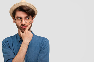 Horizontal portrait of handsome unshaven male holds chin and looks thoughtfully away, tries to plan new strategy, wears fashionable hat and jean shirt, stands against white background with blank space