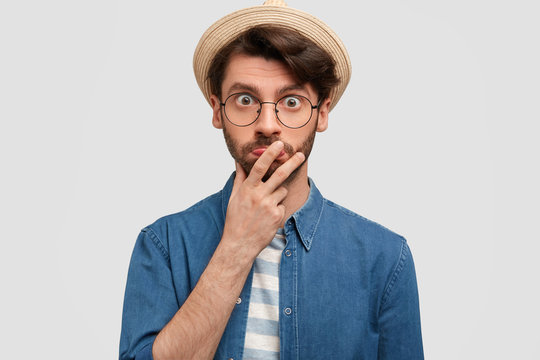 Horizontal portrait of astonished unshaven male keeps hand on mouth, looks in bewildrement, being surprised to hear news from interlocutor, wears starw hat, poses indoor against white concrete wall