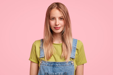 Candid shot of good looking Caucasian female with long straight hair, wears jean dungaree, looks confidently at camera, enjoys spare time, going to have walk with her dog outside. Beauty concept