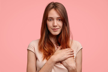 Horizontal shot of delighted friendly looking European female with freckled skin, keeps hands on heart, looks happily at camera, expresses her good feeling to boyfriend or relatives, isolated on pink