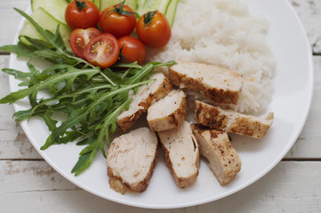 White plate with baked chicken breast, whited steamed rice, green arugula leaves (rocket salad), cucumber slices and red cherry tomatoes. Healthy eating concept