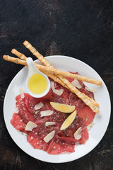 White plate with carpaccio beef over dark brown stone background, vertical shot, copy space, flatlay