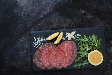 Stone slate with carpaccio beef, arugula, lemon and parmesan. View from above on a dark scratched metal background with space