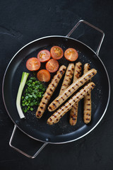 Metal pan with bbq pork sausages, tomatoes and green onion, view from above on a black stone surface, vertical shot