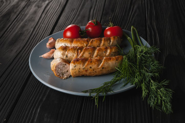 Three grilled sausages on a gray plate on a wooden black background with cherry tomatoes, herbs, garlic.