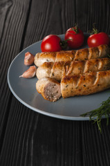 Three grilled sausages on a gray plate on a wooden black background with cherry tomatoes, herbs, garlic.