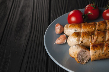 Three grilled sausages on a gray plate on a wooden black background with cherry tomatoes, herbs, garlic.
