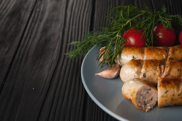 Three grilled sausages on a gray plate on a wooden black background with cherry tomatoes, herbs, garlic.