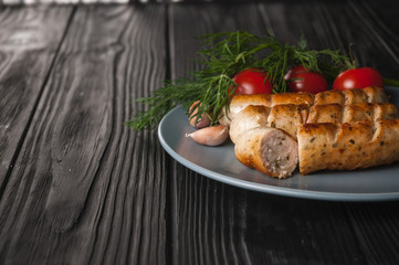 Three grilled sausages on a gray plate on a wooden black background with cherry tomatoes, herbs, garlic.
