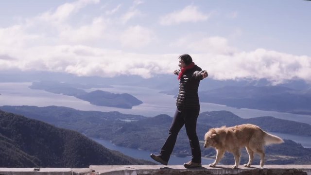 Woman with arms outstretched walking with dog over the edge of gazebo, panoramic view of Lake Nahuel Huapi. Slow motion