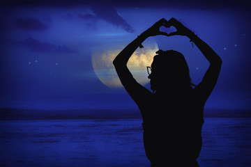 Silhouette of a girl at moonlit sky with stars near ocean holding heart - shape sign with hands. 