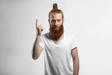 Isolated studio shot of fashionable handsome cheerful young hipster with ginger thick beard and hair knot smiling, pointing index finger upwards, indicating blank copyspace wall, promoting goods