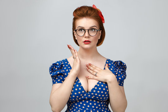 Picture of fashionable glamorous young woman wearing elegant dress, vintage hairstyle and stylish spectacles looking up, holding hand on her chest, having worried frustrated facial expression