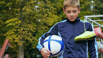 Naklejka premium Smiling teenage boy with a soccer ball in his hand and soccer boots on the shoulder against the background of the stadium. Sports training in the field.
