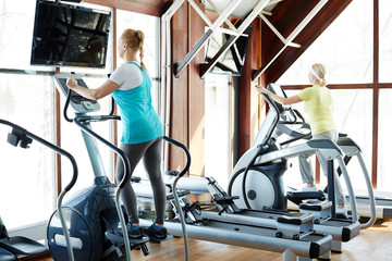 Obraz premium Two sporty mature females in activewear exercising on training equipment in modern gym