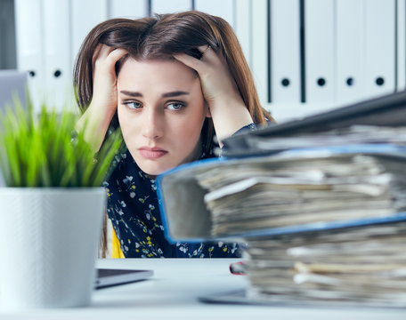 Tired and exhausted woman in spectacles looks at the mountain of documents propping up her head with her hands.