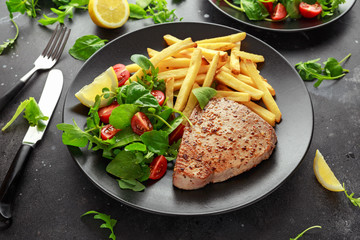 Fried Tuna Steaks on Black Plate with Fresh Green, Tomato Salad, lemon and french fries. healthy sea food
