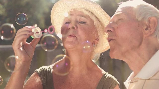 Couple of cheerful seniors outdoors. Close up elderly man and woman blowing soap bubbles together on summer nature background, slow motion. Togetherness and family values.