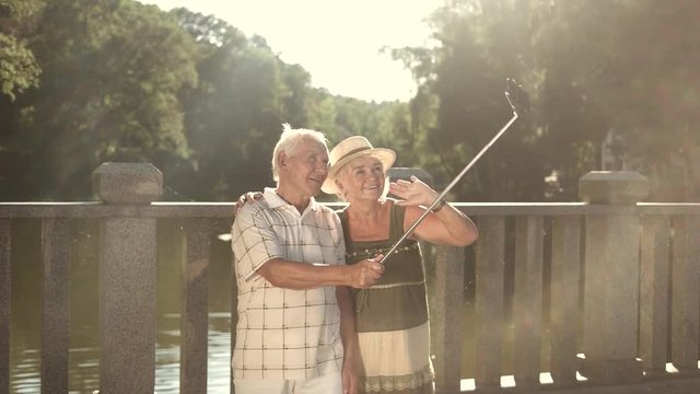 Cute couple of seniors taking a selfie. Elderly man and woman with monopod on nature background in sunny day. Old people and gadget.