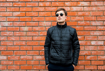 a young man stands leaning against a brick wall in sunglasses cross his arms